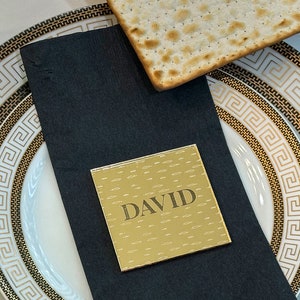 May include: A gold place card with the name "DAVID" printed on it. The place card is on a black napkin, which is on top of a white plate with a gold and black geometric pattern.