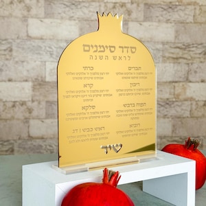 May include: A gold-colored acrylic pomegranate-shaped sign with Hebrew text, displayed on a white stand. The sign features a list of blessings for Rosh Hashanah. Two red pomegranates are visible in the image.