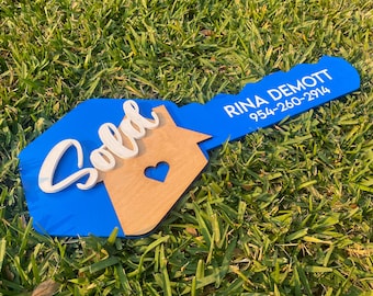May include: A blue and wood key-shaped sign with the word "Sold" in white lettering and a house outline with a heart cutout. The sign also includes the name "Rina Demott" and a phone number, "954-260-2914".