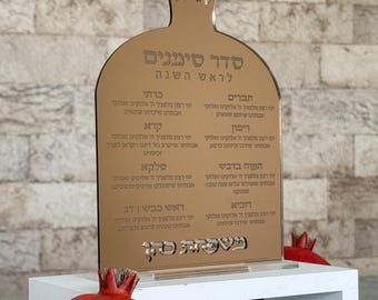May include: A gold-colored, pomegranate-shaped sign with Hebrew text, likely for Rosh Hashanah. The sign is on a white surface, with a red pomegranate visible on the right side. The sign has a decorative crown at the top.