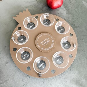 May include: A tan-colored, pomegranate-shaped plate with eight small glass bowls arranged around a central circle. Each bowl contains a silver disc with Hebrew text. A red pomegranate sits in the background.
