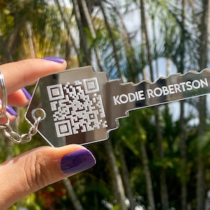 May include: A key-shaped keychain with a QR code and the name "KODIE ROBERTSON" engraved on it. The keychain is silver-colored and attached to a silver keyring. The background is blurred, showing green foliage.