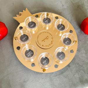 May include: A gold-colored, pomegranate-shaped serving tray with eight small glass cups. The tray has a decorative design in the center and small pomegranate cutouts around the edge. Two red pomegranates are on either side of the tray.