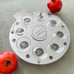 May include: A silver-colored seder plate in the shape of a pomegranate, with seven small glass bowls. The plate has Hebrew text in the center and is decorated with pomegranate shapes. Two red pomegranates are next to the plate.