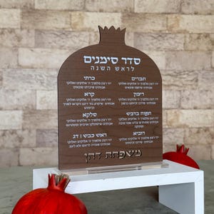 May include: A wooden sign with a pomegranate-shaped top, featuring Hebrew text and a list of symbolic foods for Rosh Hashanah. The sign is brown with a white base, and a red pomegranate is visible in the foreground.