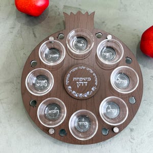 May include: A wooden pomegranate-shaped serving tray with eight clear glass bowls and a central circular design. The tray is brown with silver accents and includes text in Hebrew. Two red pomegranates are visible.