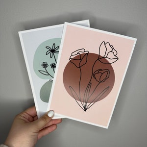 Può includere: Due stampe d'arte stampabili con disegni di fiori minimalisti a linee. La prima stampa ha uno sfondo verde con un solo fiore al centro. La seconda stampa ha uno sfondo rosa con tre fiori al centro.