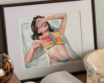May include: A framed watercolor illustration depicts a person lying down with a book titled Beach Read covering their face. The artwork is displayed alongside an open book, a glass of iced coffee, a textured bowl, and a brown patterned glass object.