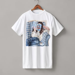 Puede incluir: Camiseta blanca con una ilustración en acuarela de una persona leyendo un libro en un sillón a cuadros azules. El título del libro es visible. Una taza de café está sobre una mesita. La camiseta está colgada en una percha de madera.