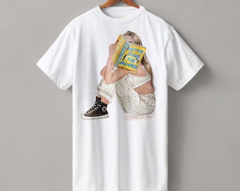 May include: A white short-sleeve t-shirt featuring a graphic of a person sitting and reading a yellow book titled Better Than The Movies by Lynn Painter. The person wears striped pants and black high-top sneakers while holding the book up.