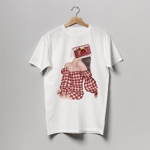 May include: White t-shirt with a graphic print of a woman wearing a red and white gingham dress. The woman's head is covered by a box with the text "CHERRY" on it.