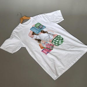 Puede incluir: Camiseta blanca con una ilustración en acuarela de una persona tumbada con un libro sobre la cara. El diseño incluye otros libros y una almohada a cuadros verdes y blancos. La camiseta está colgada en una percha de madera.