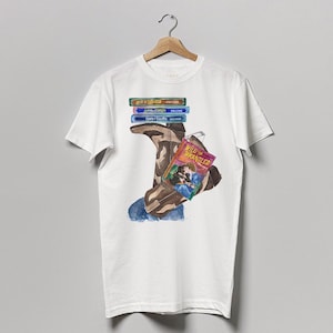 Puede incluir: Camiseta blanca con una ilustración de dibujos animados de una persona que lleva una bota marrón y sostiene un libro titulado "Wild & Wrangled". La persona sostiene una pila de cintas VHS de colores.