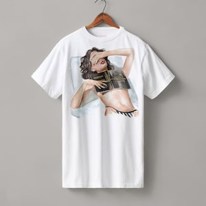 May include: A white short-sleeve t-shirt featuring a graphic illustration of a person with dark hair lying down, covering their eyes with one hand while holding a black book titled ALCHEMISED over their chest, wearing black and white striped bottoms.