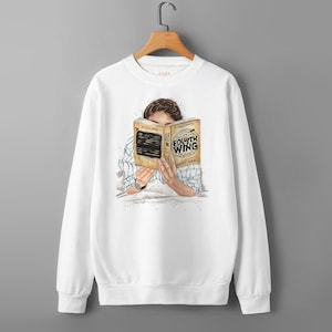 May include: White sweatshirt with an illustration of a person reading a book titled "Fourth Wing" by Rebecca Yarros. The book is held up to the person's face. The sweatshirt is hanging on a wooden hanger.