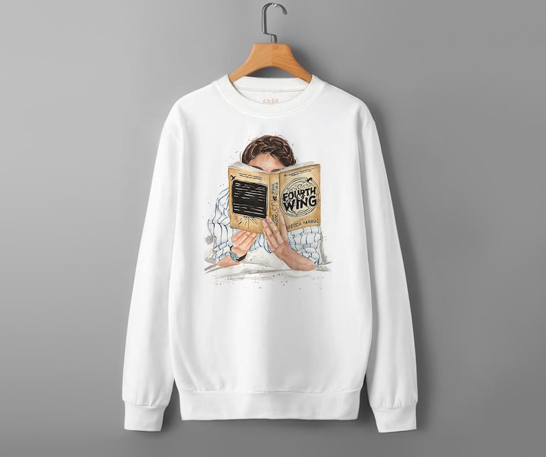 May include: White sweatshirt featuring an illustration of a person reading a book titled "Fourth Wing" by Rebecca Yarros. The book is held up to the person's face. The sweatshirt is hanging on a wooden hanger.