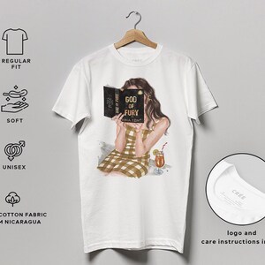 May include: White unisex t-shirt featuring an illustration of a person reading a book titled "GOD OF FURY." The person is wearing a brown and tan plaid dress and holding a cocktail. The shirt is made of 100% cotton fabric from Nicaragua.