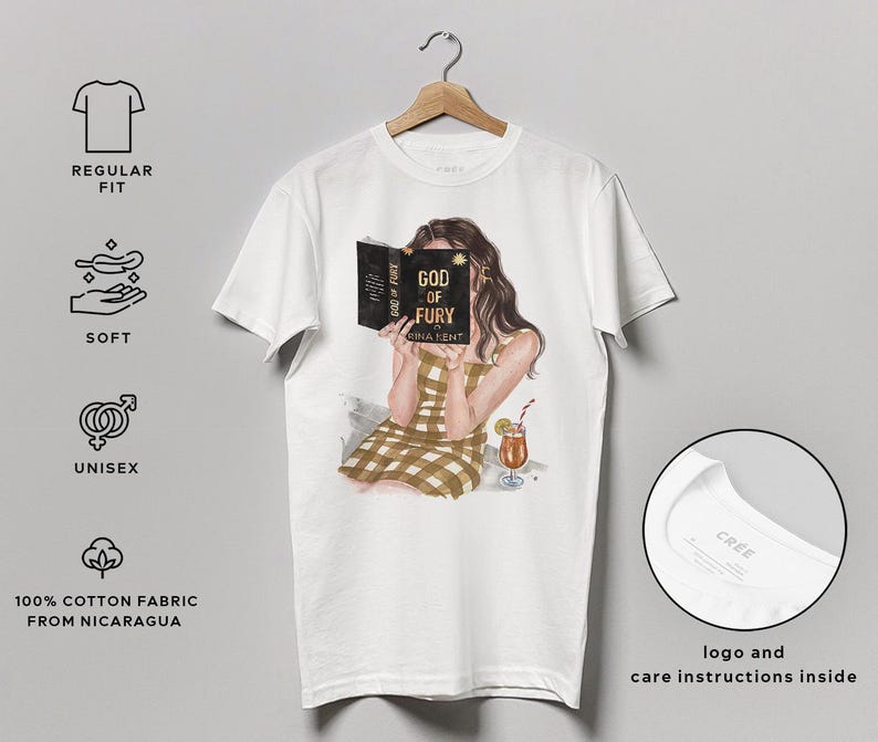 May include: White unisex t-shirt featuring an illustration of a person reading a book titled "GOD OF FURY." The person is wearing a brown and tan plaid dress and holding a cocktail. The shirt is made of 100% cotton fabric from Nicaragua.