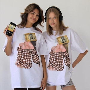 Puede incluir: Dos camisetas blancas con un estampado gráfico de una persona con el libro Fourth Wing de Rebecca Yarros como cabeza, vistiendo un top de hombros descubiertos a cuadros marrones y blancos. El texto dice: Both models are wearing size L for an oversized fit.