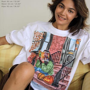 Peut inclure: Un t-shirt blanc à manches courtes avec une illustration graphique colorée représentant une personne assise sur des marches en briques lisant un livre intitulé It's Different This Time, entourée de feuilles d'automne et de deux citrouilles.