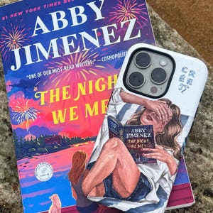 May include: A paperback book titled The Night We Met by Abby Jimenez featuring a colorful fireworks illustration, positioned beneath a white smartphone case decorated with an artistic graphic of a person reading the same book while lying down.