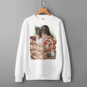May include: White sweatshirt featuring an illustration of a person reading a book titled "ALCHEMIED" while wrapped in a plaid blanket. A vase of autumn leaves sits nearby. The design has a cozy, fall theme.