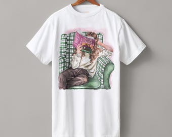 May include: White t-shirt featuring an illustration of a person reading a book while seated in a green plaid armchair. The book's cover is visible, with the title "Planet Own League". The person is wearing a white sweater and brown pants.