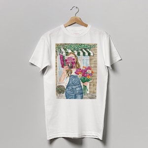 Op de afbeelding: Wit T-shirt met een afbeelding van een vrouw die een boek met de titel "Practice Makes Perfect" en een boeket roze en paarse bloemen vasthoudt. De vrouw staat voor een gebouw met een groene luifel.