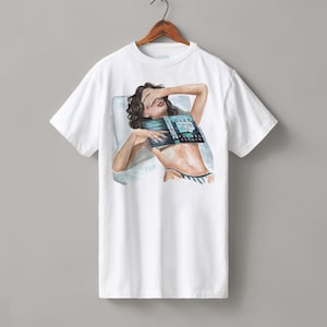 Könnte beinhalten: Weißes T-Shirt mit einer Illustration einer Person, die liegt und ein Buch vor ihrem Gesicht hält. Der Titel des Buches lautet "DARK SECRETS". Die Person trägt schwarz-weiß gestreifte Unterwäsche. Das T-Shirt hängt an einem Holzkleiderbügel.