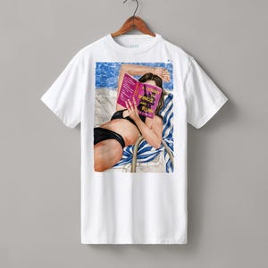 Könnte beinhalten: Weißes T-Shirt mit einer Aquarellillustration einer Person, die ein Buch mit dem Titel "Court of Wings and Ruin" liest, während sie sich sonnt. Die Illustration zeigt einen schwarzen Bikini und einen blau-weiß gestreiften Liegestuhl.