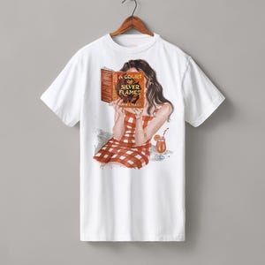 May include: A white short-sleeve t-shirt featuring a graphic illustration of a person with long dark hair wearing a red and white checkered dress. The person is holding a book titled A Court of Silver Flames by Sarah J. Maas and a drink.