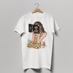 May include: White t-shirt with a graphic of a person reading a book titled "GOD OF FURY". The person is wearing a plaid dress and has a drink with a straw next to them. The shirt is hanging on a wooden hanger.