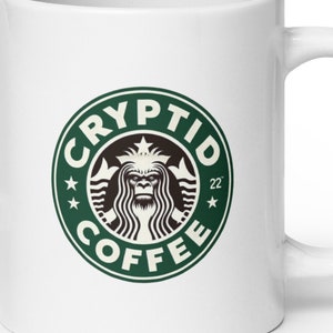 May include: White ceramic coffee mug with a green and brown Cryptid Coffee logo. The logo features a Bigfoot-like creature in the center, surrounded by text. The mug has a curved handle and is designed for hot beverages.