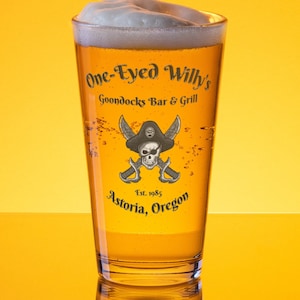 Pode incluir: Um copo alto de cerveja com uma cabeça branca. O copo tem um logotipo que diz "One-Eyed Willy's Goondocks Bar & Grill Est. 1985 Astoria, Oregon" com uma caveira de pirata e ossos cruzados.