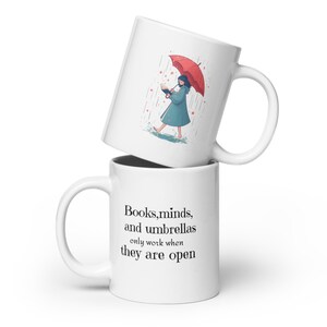Puede incluir: Taza de cerámica blanca con una cita en negro que dice "Los libros, las mentes y los paraguas solo funcionan cuando están abiertos". La taza está apilada sobre otra taza de cerámica blanca con una ilustración de dibujos animados de una persona caminando bajo la lluvia con un paraguas y leyendo un libro.
