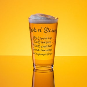 Könnte beinhalten: Ein hohes Glas gefüllt mit einem hell bernsteinfarbenen Getränk, das mit einer dicken Schicht weißen Schaums gekrönt ist. Das Glas trägt ein gedrucktes Rezept für einen "Dark 'n' Stormy" Cocktail, auf dem die Zutaten aufgeführt sind: 30 ml gewürzter Rum, 10 ml Limettensaft, 150 ml Ginger Beer und eine Garnitur aus Limettenspalten und kandiertem Ingwer.
