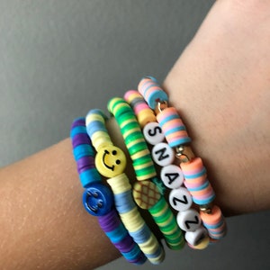 May include: A stack of colorful beaded bracelets with smiley face and pineapple charms. The bracelets spell out the name "Snazzy" in black letters.