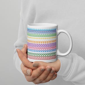 May include: White ceramic mug featuring a colorful, repeating pattern of stitched designs in rainbow hues. The design includes various shapes like arches, zigzags, and circles. The mug has a curved handle and is held by a person.