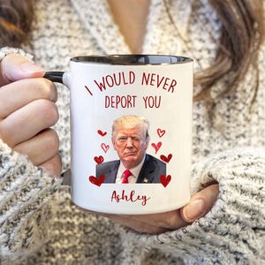 May include: White ceramic coffee mug with a black rim and handle. The mug features a red heart design with the text "I would never deport you" and a photo of a man with the name "Ashley" printed below.