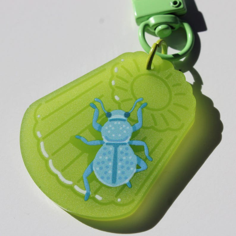 May include: A lime green keychain with a light blue insect design. The keychain has a rectangular shape with a scalloped edge and a sun graphic. The insect has a dotted pattern and is attached to a green clasp.