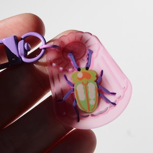 May include: A colorful keychain featuring a cartoon beetle design. The beetle has a green and orange body with blue legs and antennae. The keychain is attached to a purple clasp and a pink, flower-shaped charm.