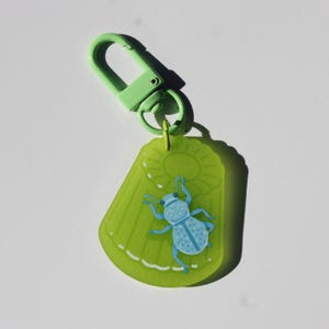 May include: A green keychain with a light green clip. The keychain has a lime green, jelly-like shape with a blue beetle illustration. The beetle has a dotted pattern and a hat. The keychain is a fun accessory.