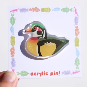 May include: A colorful acrylic pin featuring a wood duck design. The duck has a green head, orange beak, and yellow and black body. The pin is on a white card with a border of insect illustrations and the text "acrylic pin!"