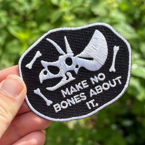 May include: Black and white embroidered patch featuring a Triceratops skull and bones design. The patch has the text "MAKE NO BONES ABOUT IT." The patch is an oval shape with a black background and white stitching.