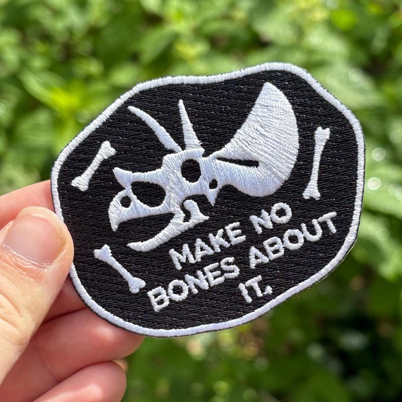 May include: Black and white embroidered patch featuring a Triceratops skull and bones design. The patch has the text "MAKE NO BONES ABOUT IT." The patch is an oval shape with a black background and white stitching.