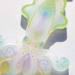 May include: A whimsical, translucent octopus-shaped ornament. The design features pastel colors, including light green, yellow, and purple, with delicate dotted and swirled details. The octopus has a stylized appearance, with a playful and artistic aesthetic.