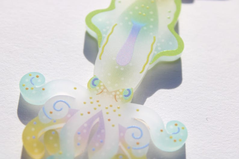 May include: A whimsical, translucent octopus-shaped ornament. The design features pastel colors, including light green, yellow, and purple, with delicate dotted and swirled details. The octopus has a stylized appearance, with a playful and artistic aesthetic.