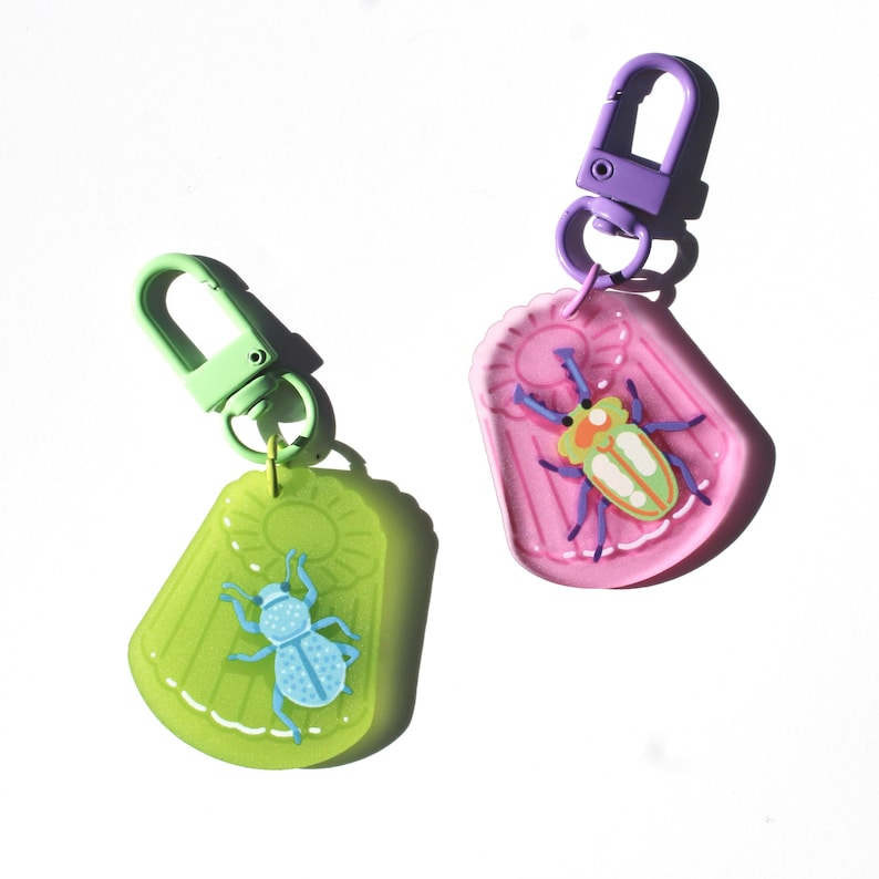 May include: Two keychains with insect designs. One is lime green with a blue bug, the other is pink with a yellow and green bug. Both have a sun design and a metal clasp in green and purple.