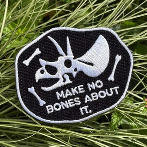 May include: Black and white embroidered patch featuring a Triceratops skull and bones. The patch has the text "MAKE NO BONES ABOUT IT" in white lettering. The patch is surrounded by a black border.