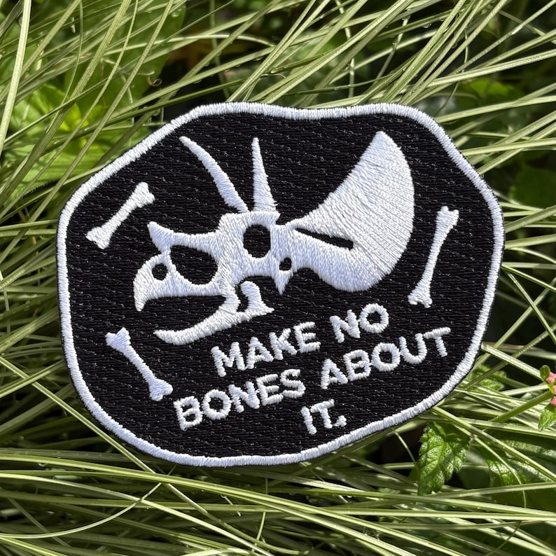 May include: Black and white embroidered patch featuring a Triceratops skull and bones. The patch has the text "MAKE NO BONES ABOUT IT" in white lettering. The patch is surrounded by a black border.